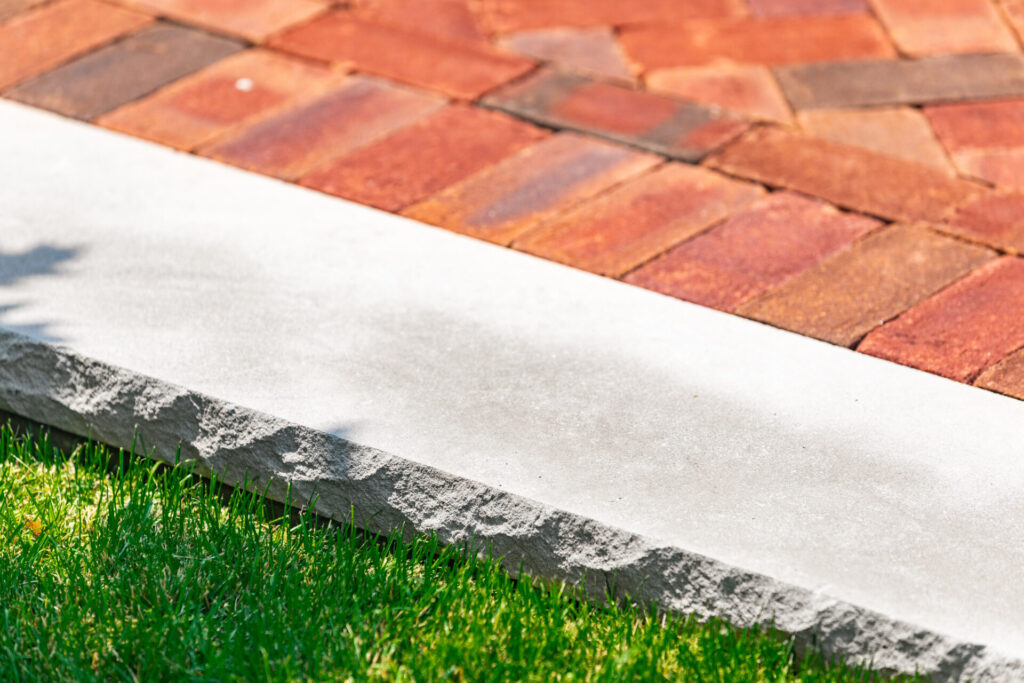 This image shows a close-up of a neatly mowed green lawn, a white concrete curb, and a section of red brick paving under bright sunlight.