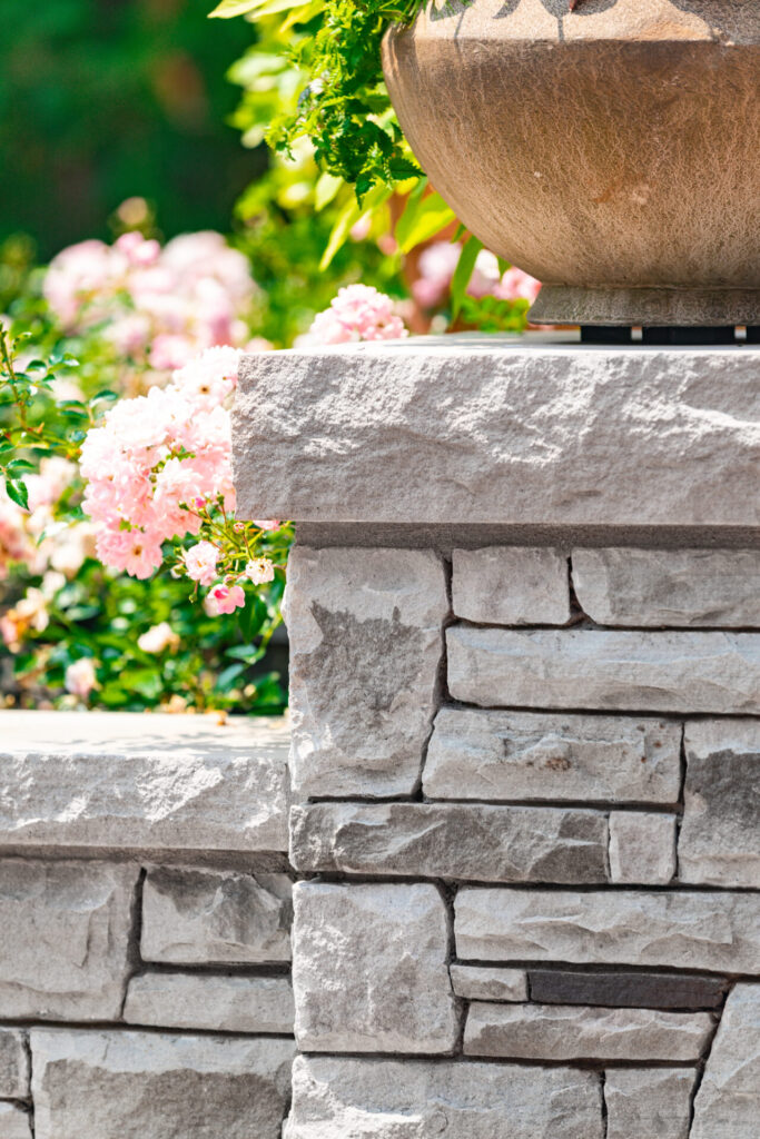 This image shows a partial view of a textured stone pillar with a decorative vase on top, against a blurred background of pink flowers and greenery.