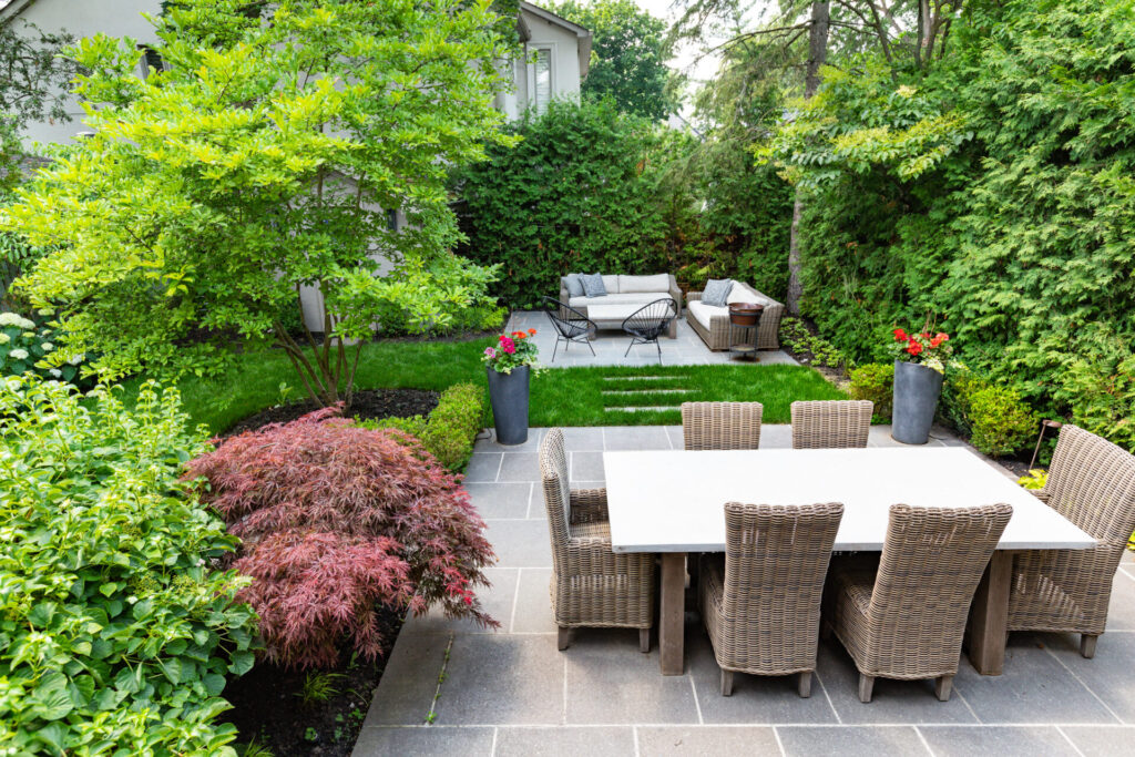 This is a well-manicured garden with a dining area, wicker chairs, a lush lawn, diverse plants, and a cozy lounging spot amidst verdant greenery.