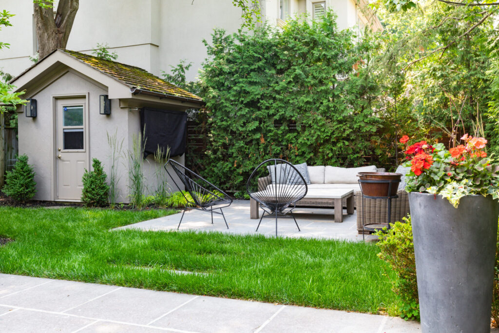 A serene backyard with lush green grass features an outdoor seating area, modern black chair, shed, plants, and a blooming flower pot.