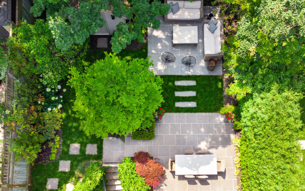 Aerial view of a landscaped garden with a stone patio, outdoor furniture, lush green trees, and well-maintained hedges, alongside a clear pathway.