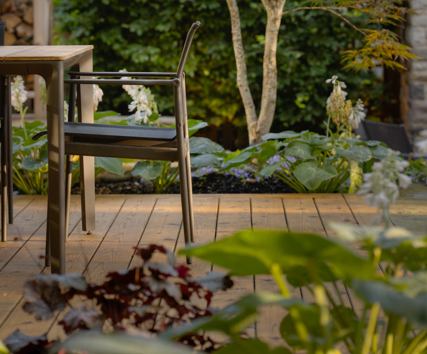 A tranquil garden patio setting with a metal chair and wooden table, surrounded by lush greenery, foliage, and a soft-focus backdrop of plants.