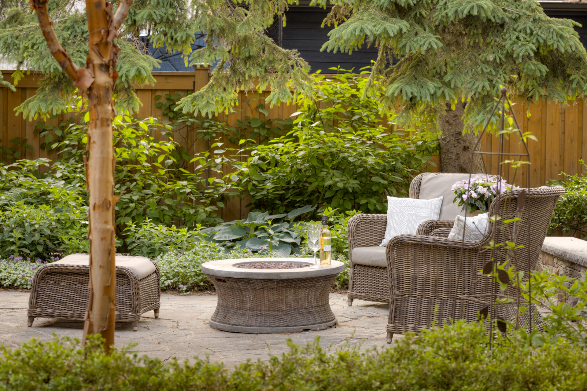 An inviting garden patio with wicker furniture, lush greenery, and a glass of champagne on the table, set against a wooden fence backdrop.