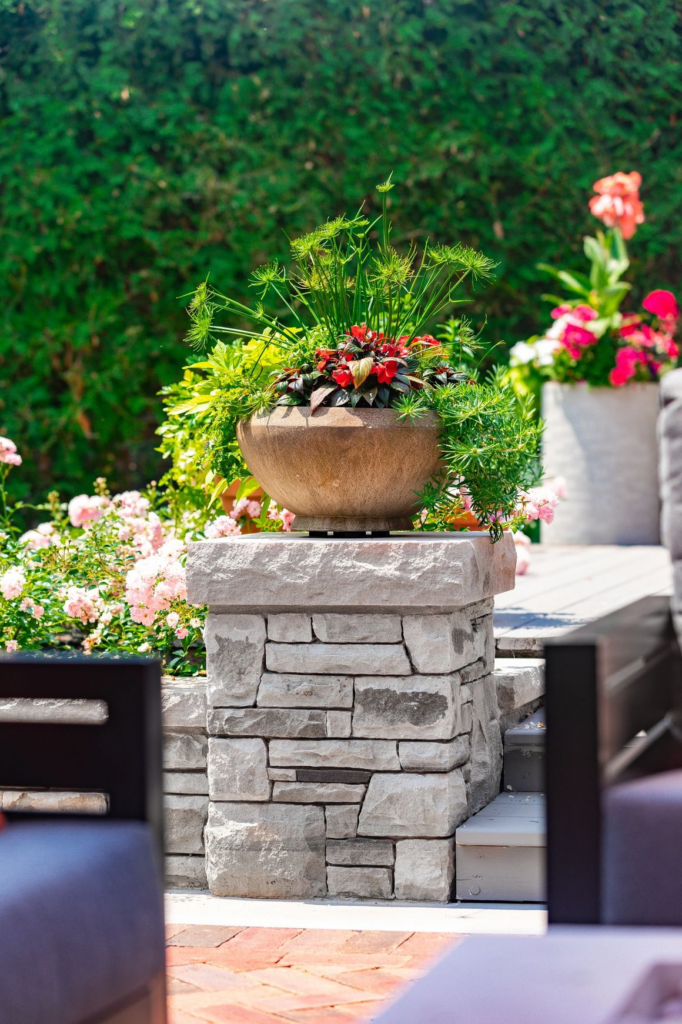 An outdoor stone planter with lush greenery and red flowers. Patio furniture in foreground. Background shows flowering shrubs under a sunny sky.