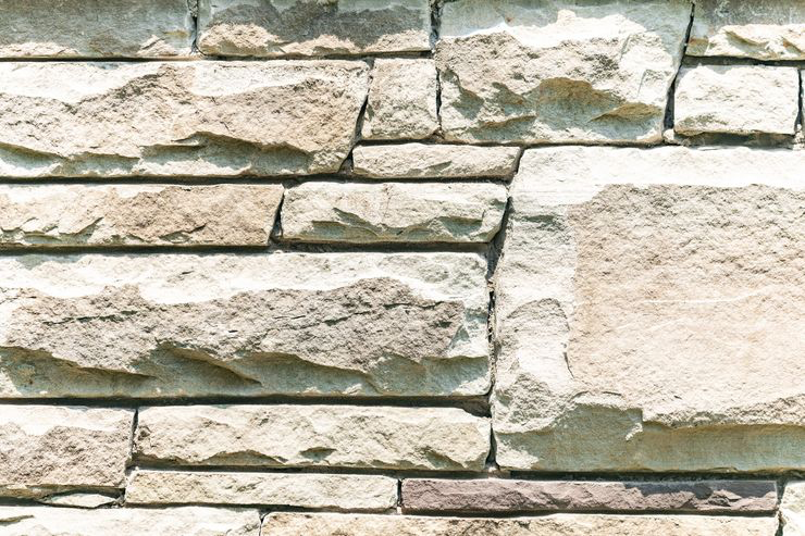 This image shows a close-up of a stone wall with irregularly shaped stones in various shades of beige, arranged in horizontal layers, under bright lighting.