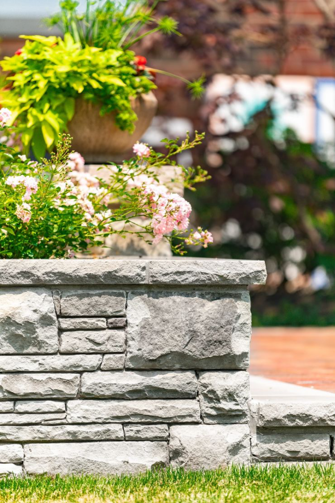 An ornate flowerpot sits atop a stone wall, overflowing with greenery and pink blossoms, set against a soft-focused background of a residential setting.