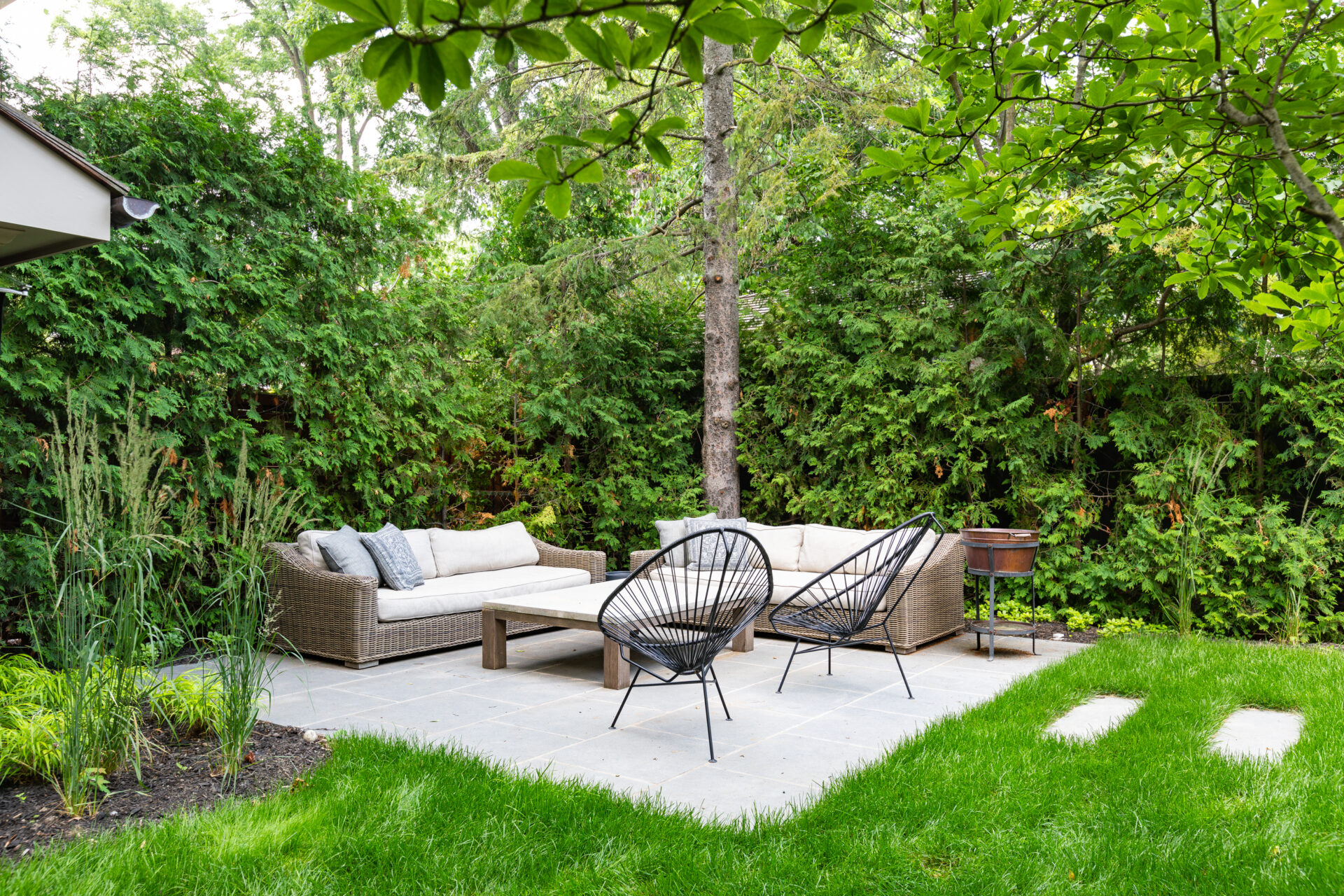 A tranquil garden patio with a wicker sofa, two black chairs, greenery all around, a lush lawn, and stepping stones in a serene setting.