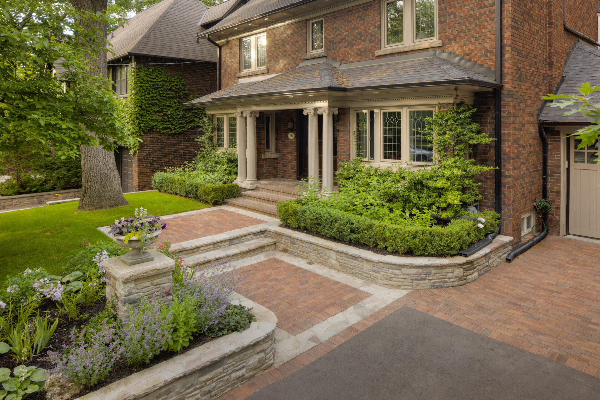 A traditional brick house with white columns, stone steps, landscaped garden, and a driveway featuring pavers and greenery under a clear sky.