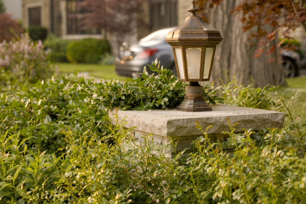 An outdoor lamp post sits atop a stone pedestal, surrounded by leafy green shrubs, with a dappled sunlight and a residential backdrop.