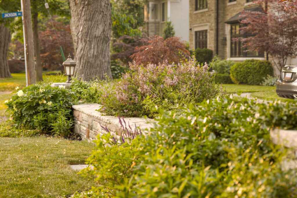 This image shows a lush, well-maintained garden in front of a house, with large trees, flowering shrubs, and a stone-bordered flowerbed under a street sign.