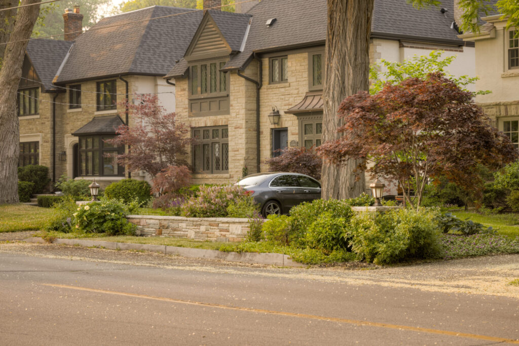 This image showcases a large, two-story house made of stone with a steeply pitched roof, surrounded by lush greenery, with a car parked in the driveway.