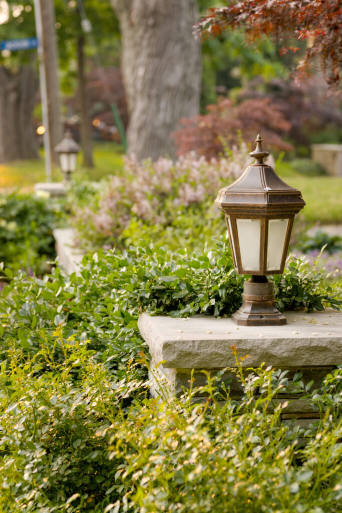 This image features an ornate outdoor lamp post on a stone edge, surrounded by lush greenery and blooming plants in a serene garden setting.