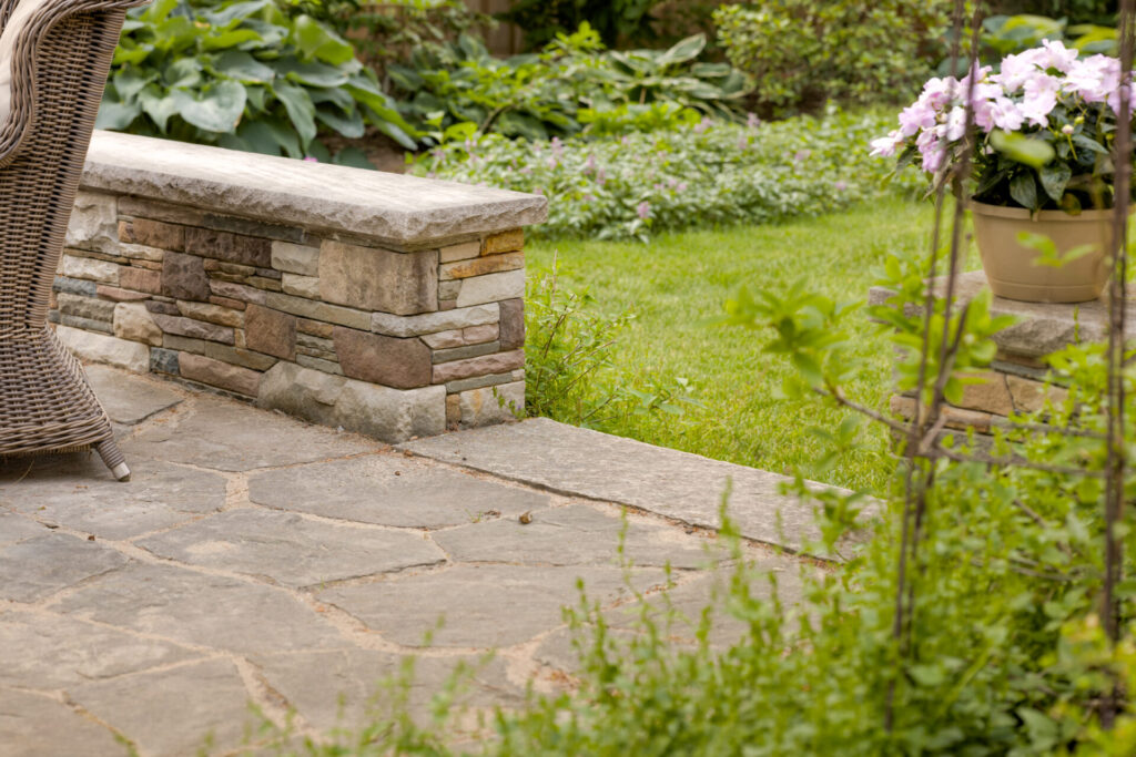 A tranquil garden scene featuring a stone bench, a wicker chair, flagstone patio, lush green lawn, flowering plants, and a serene, inviting atmosphere.