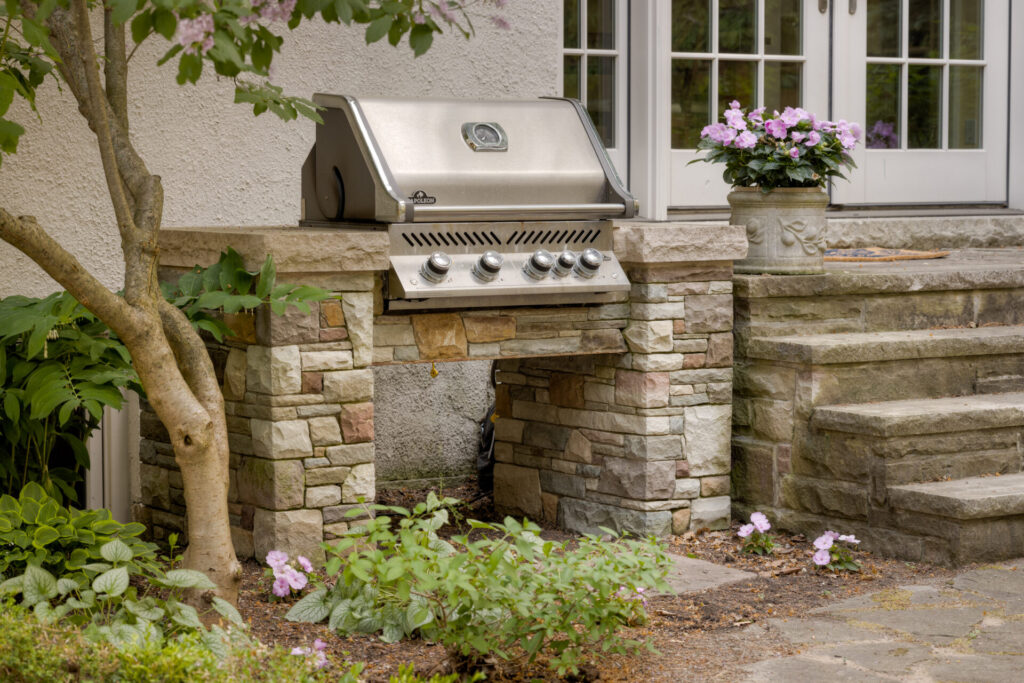 An outdoor stainless steel grill is built into a stone island next to steps leading to a house's entrance, surrounded by plants and flowers.