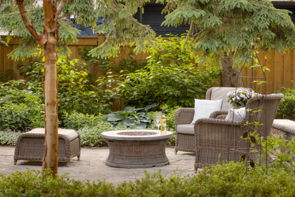 Cozy garden patio with wicker furniture, including chairs, a footrest, and a table with a glass and a vase with flowers, surrounded by green plants.