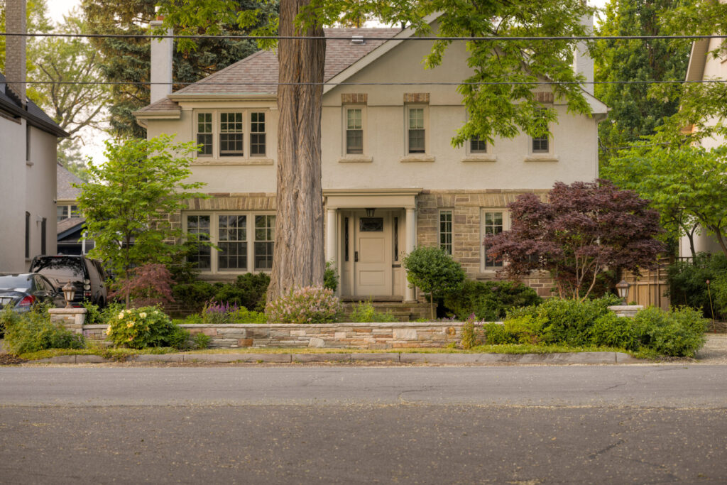 A two-story suburban house with beige siding, stone accents, landscaped front yard, steps leading to a doorway, and a car parked on the side.