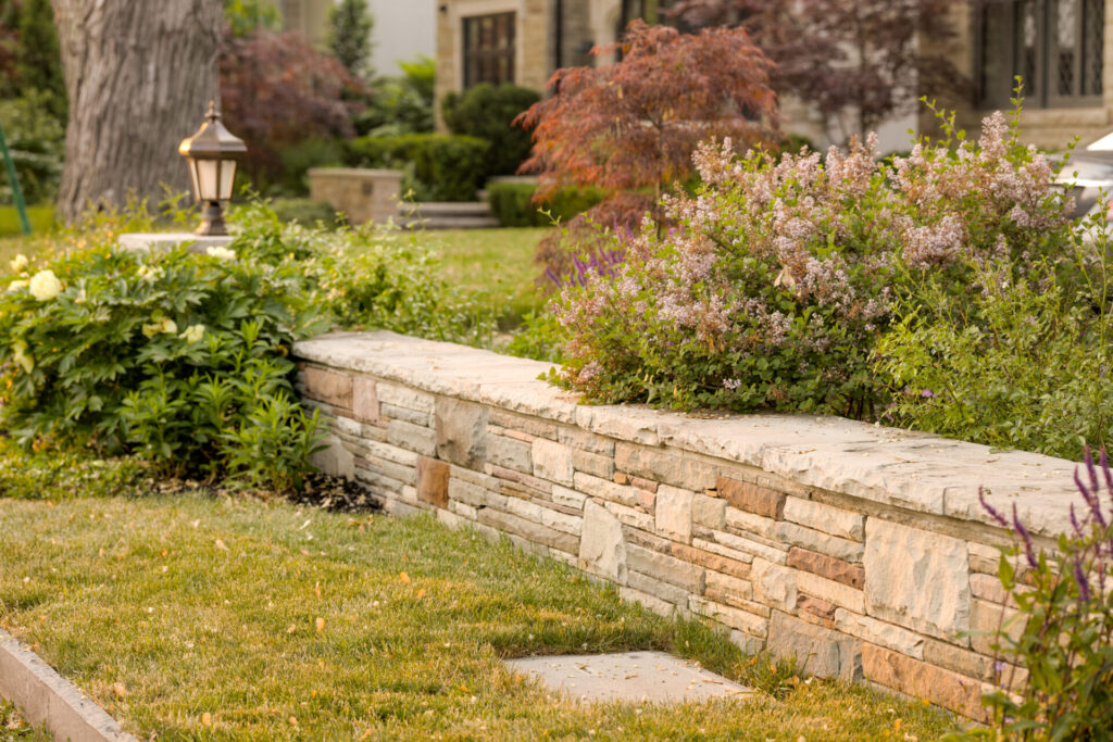 This image shows a neatly landscaped garden with a stone retaining wall, flowering shrubs, a lamp post, and a well-maintained lawn in daylight.