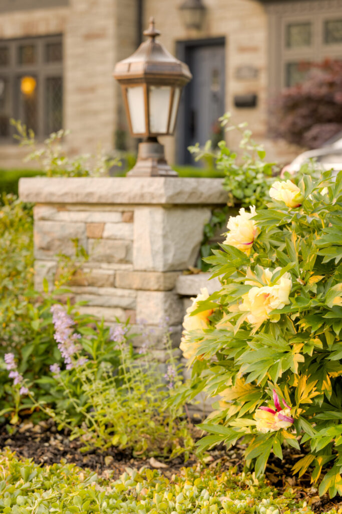 An outdoor lamp post stands atop a stone pillar amidst vibrant yellow flowers and greenery with a blurred background of an elegant building.