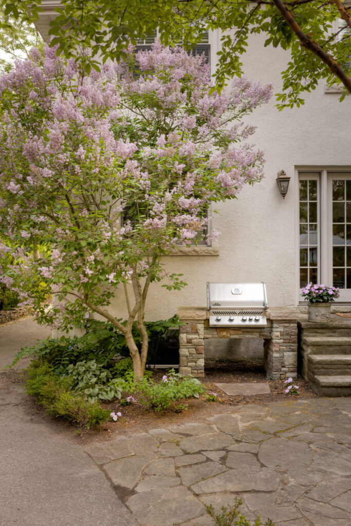 A tree with pink blossoms dominates the foreground in front of a house with stucco walls and a built-in barbecue grill on a stone patio.