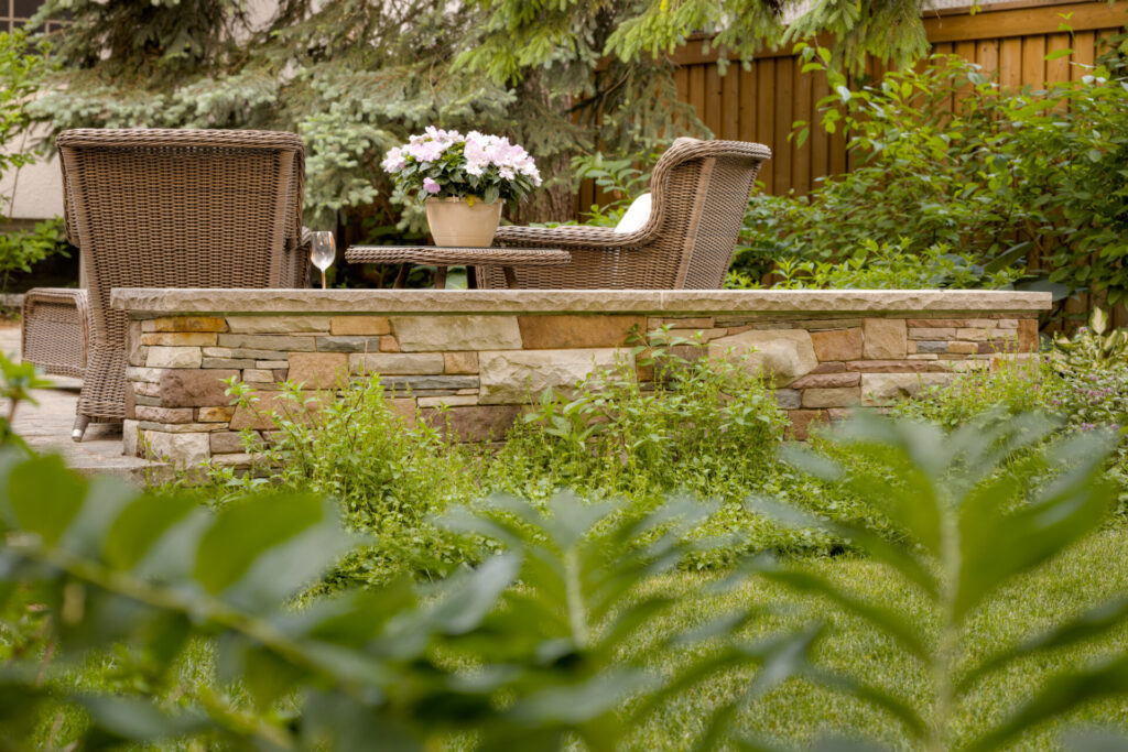 A serene garden setting with a stone bar counter, rattan chairs, and potted flowers. Foliage frames the tranquil outdoor space for relaxation.