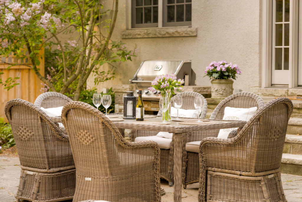 An inviting outdoor dining area with wicker chairs around a table set with wine glasses, flowers, and a bottle, adjacent to a barbecue grill.