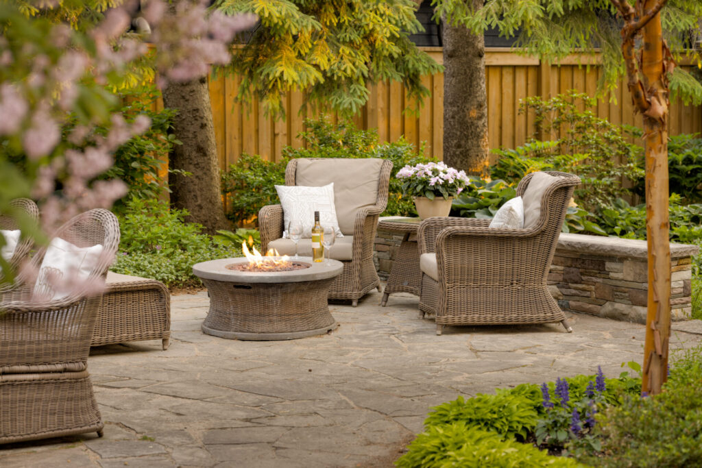 An outdoor patio with wicker furniture centered around a fire pit. There are cushions, flowering plants, and a surrounding wooden fence with greenery.