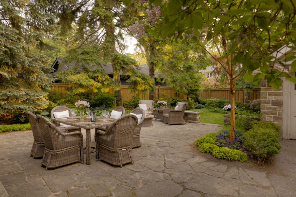 A tranquil backyard setting with wicker furniture, lush landscaping, a stone patio, and mature trees. An inviting outdoor dining area is highlighted.