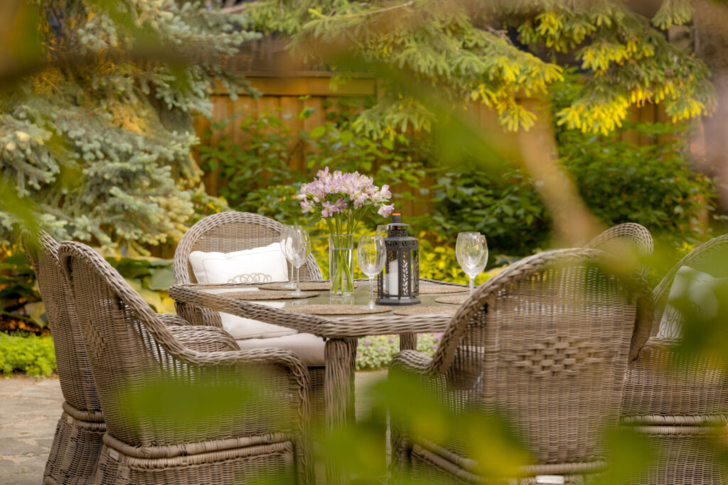 A serene garden setting with wicker chairs around a table, fresh flowers, and elegant glassware hinting at an intimate outdoor dining experience.