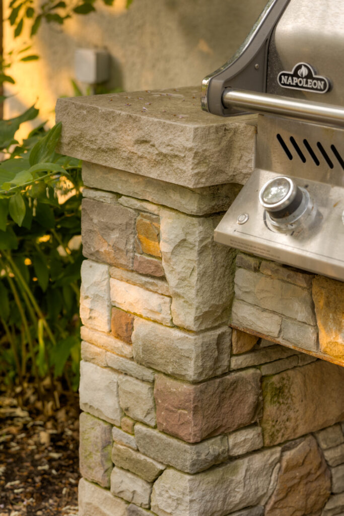 An outdoor stone pillar supports a corner of a stainless steel Napoleon grill, with foliage and a warmly lit background suggesting a garden setting.