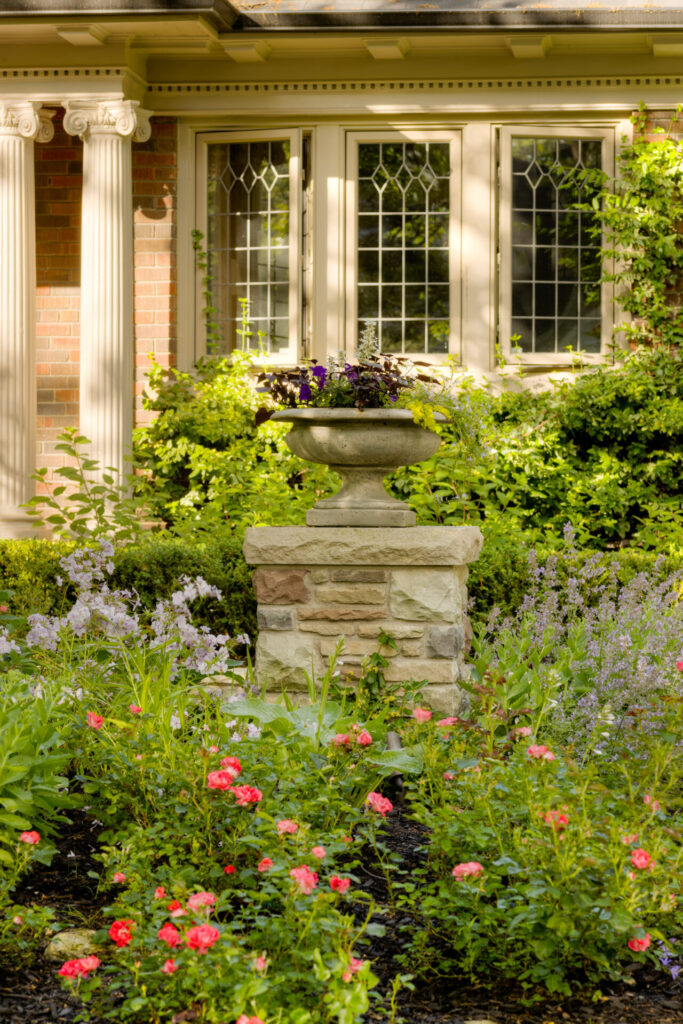 A tranquil garden with a stone pedestal planter, colorful flowers, and classic windows framed by elegant columns in a serene, residential setting.