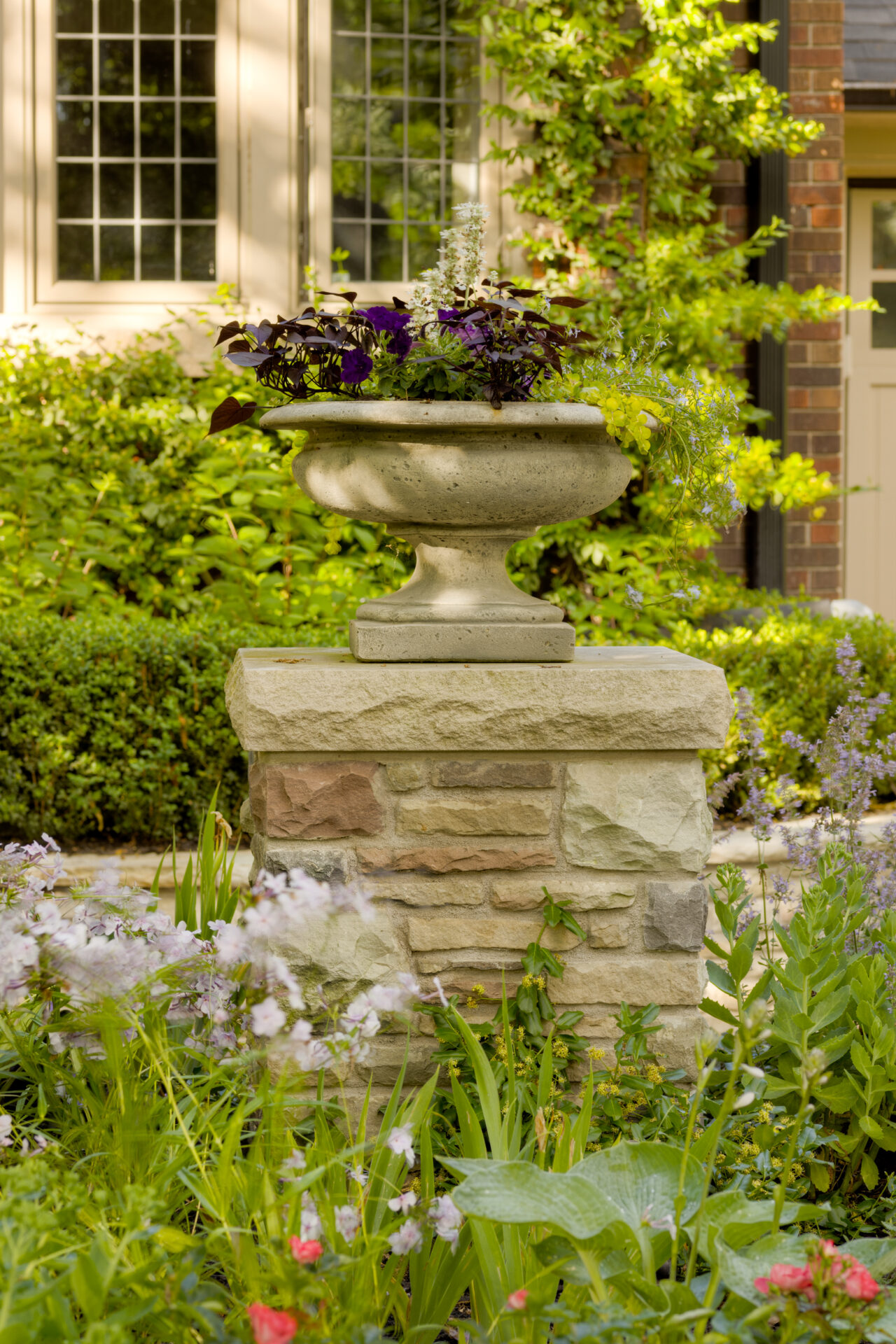 A classical stone planter with purple and green plants sits atop a column amidst a lush garden, with a brick building and window in the background.