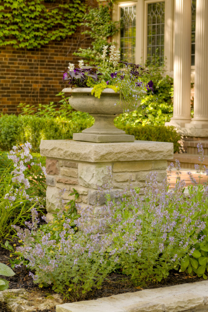 A stone urn filled with a colorful array of flowers stands atop a pedestal in a lush garden, with a brick structure and pillars in the background.