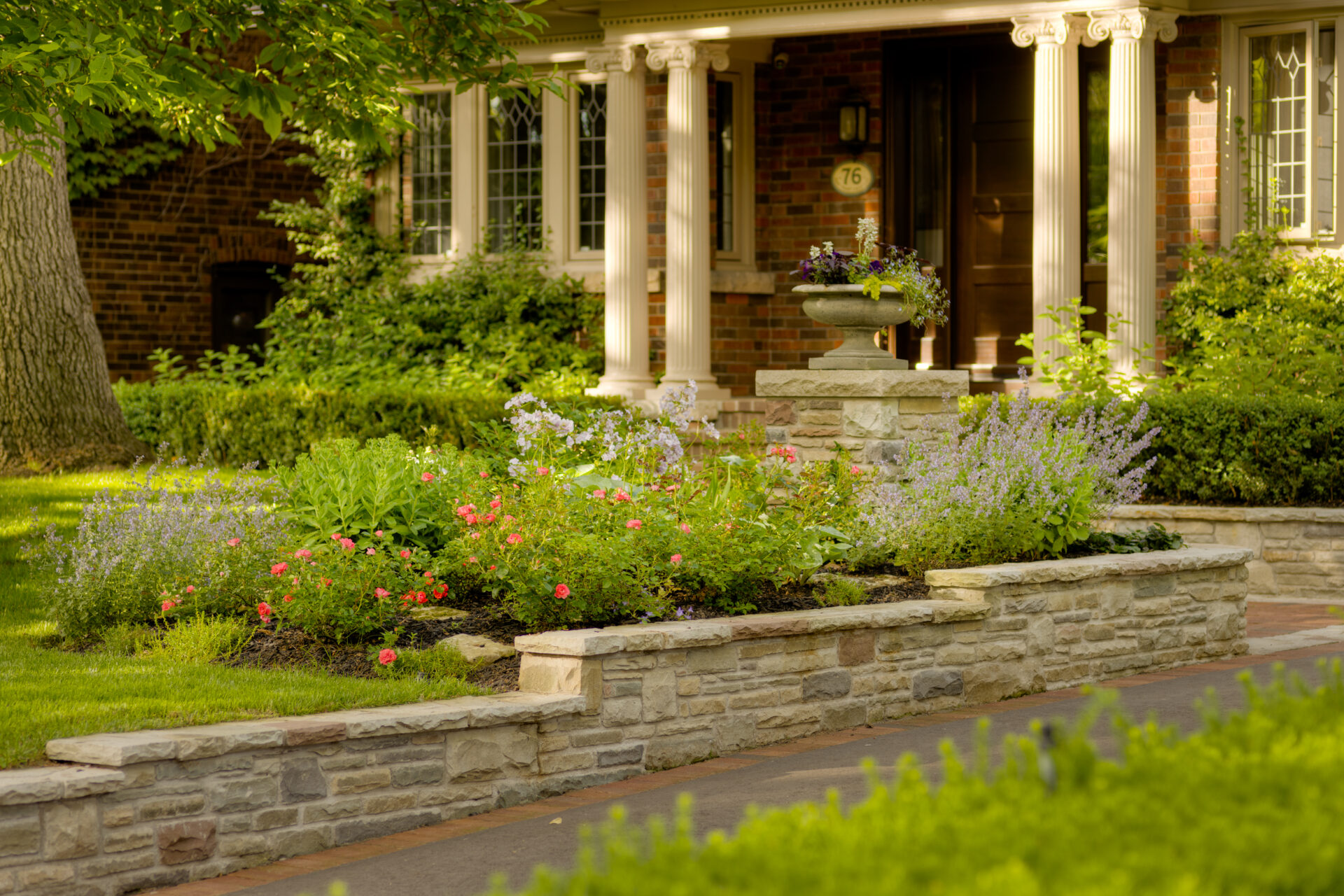 A serene front yard of a brick house with lush gardens, flowering plants, a stone pathway, and classical white columns near the entrance.