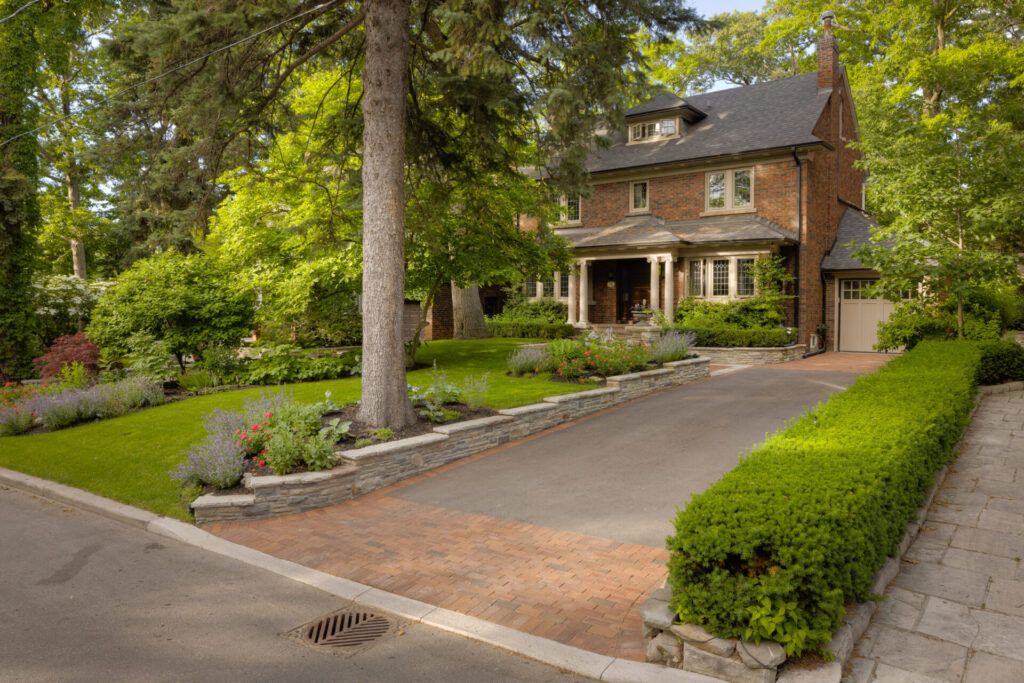 An elegant brick house with a landscaped garden, stone-bordered flower beds, lush greenery, and a curved driveway on a bright, sunny day.