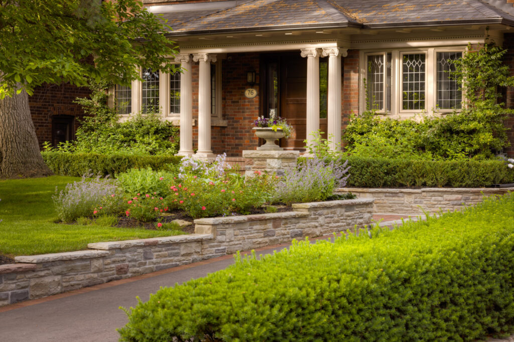 This image displays a charming house with a stone pathway, lush landscaping, classical columns, windows with grids, and flowering plants adding a welcoming touch.