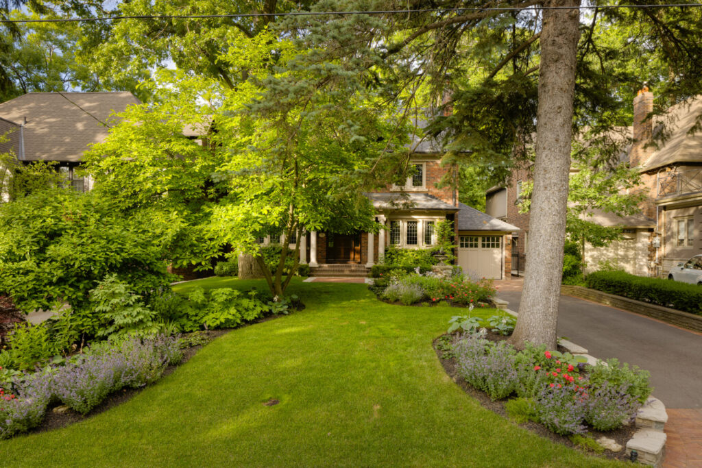 The image shows a lush, landscaped front yard with flowering plants and a well-maintained lawn leading to a traditional brick house, partially obscured by trees.