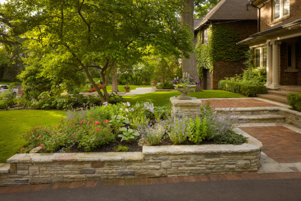 A well-manicured garden with vibrant flowers in front of an elegant brick house, featuring a stone pathway leading to a porch and lush greenery.