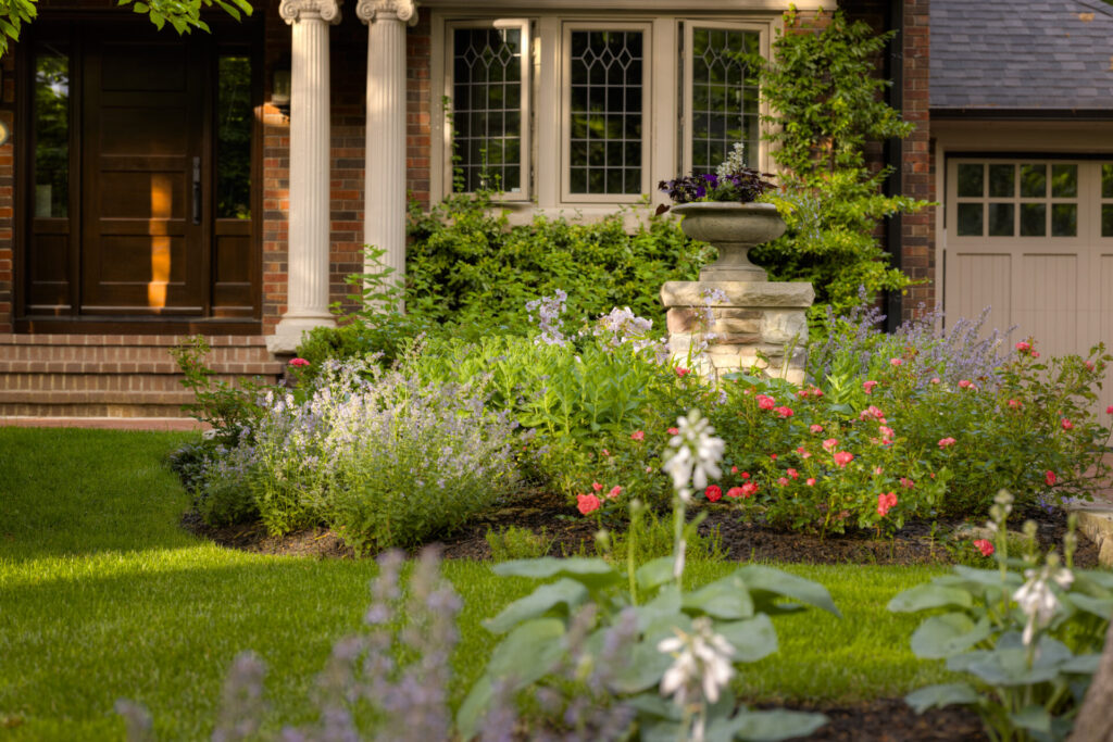 A serene and well-maintained garden with flowering plants in front of an elegant brick house with white columns and a classical entryway.