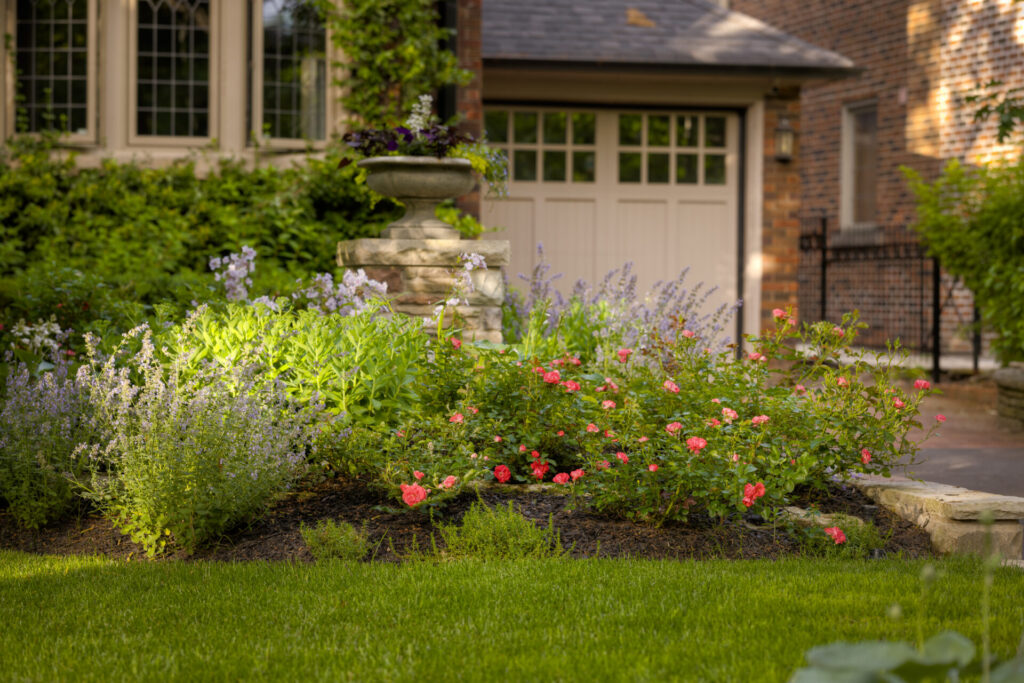 A well-manicured garden with various flowers in front of a traditional house with large windows, a stone urn, and a neatly trimmed lawn.