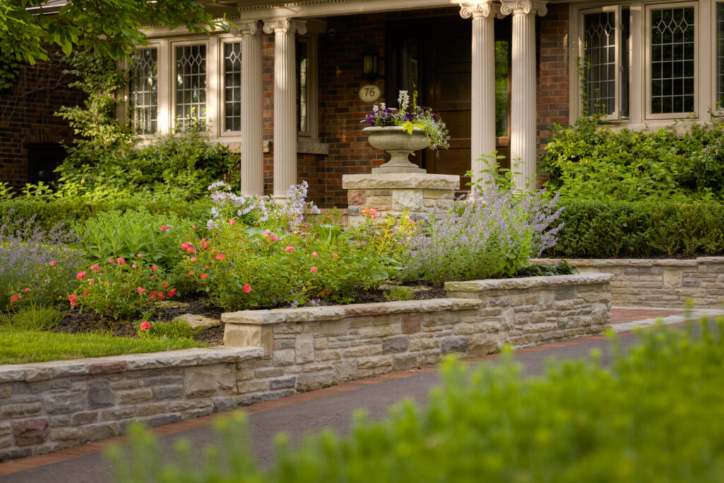 The image shows a well-manicured front yard garden with colorful flowers, stone walls, a birdbath, and a classical-style home with elegant columns.