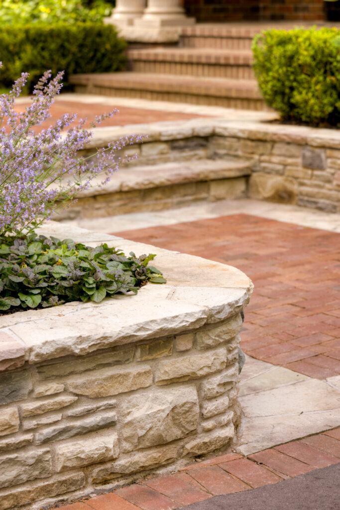 Natural stone steps lead up to a brick building. Plants grow alongside the staircase, adding a touch of greenery and purple flowers to the scene.