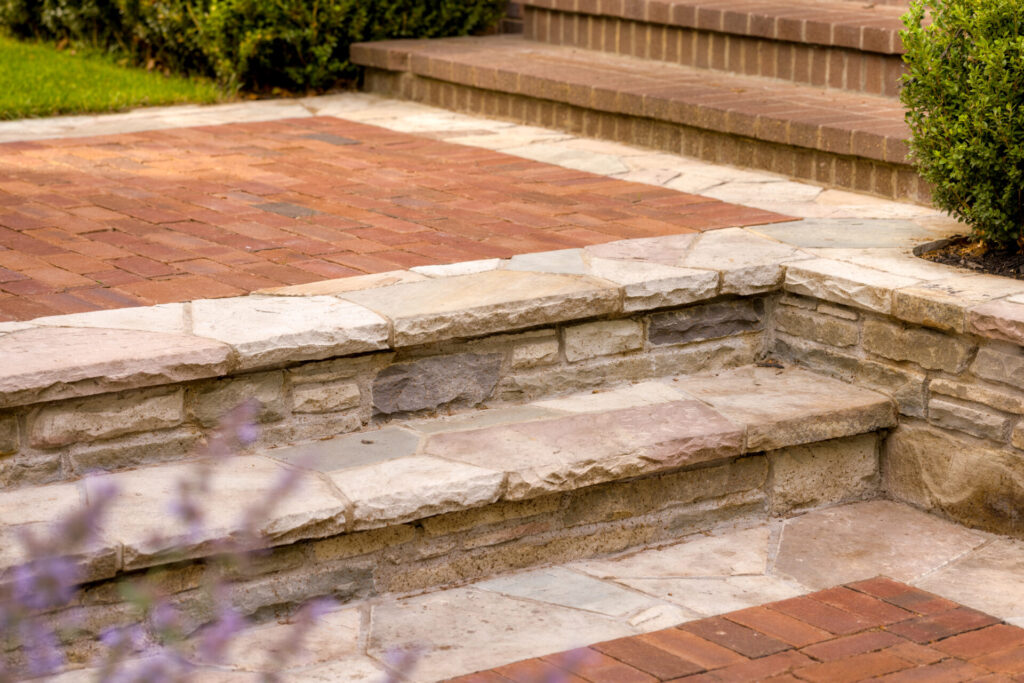 The image shows a series of brick and stone steps and a paved area leading up to a garden with a stone retaining wall and green shrubbery.