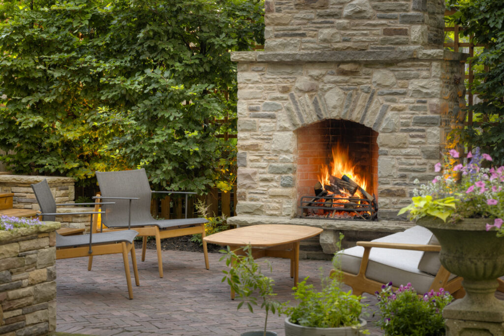 A cozy backyard patio with a stone fireplace ablaze, surrounded by lush greenery, comfortable modern furniture, and vibrant flowering plants.