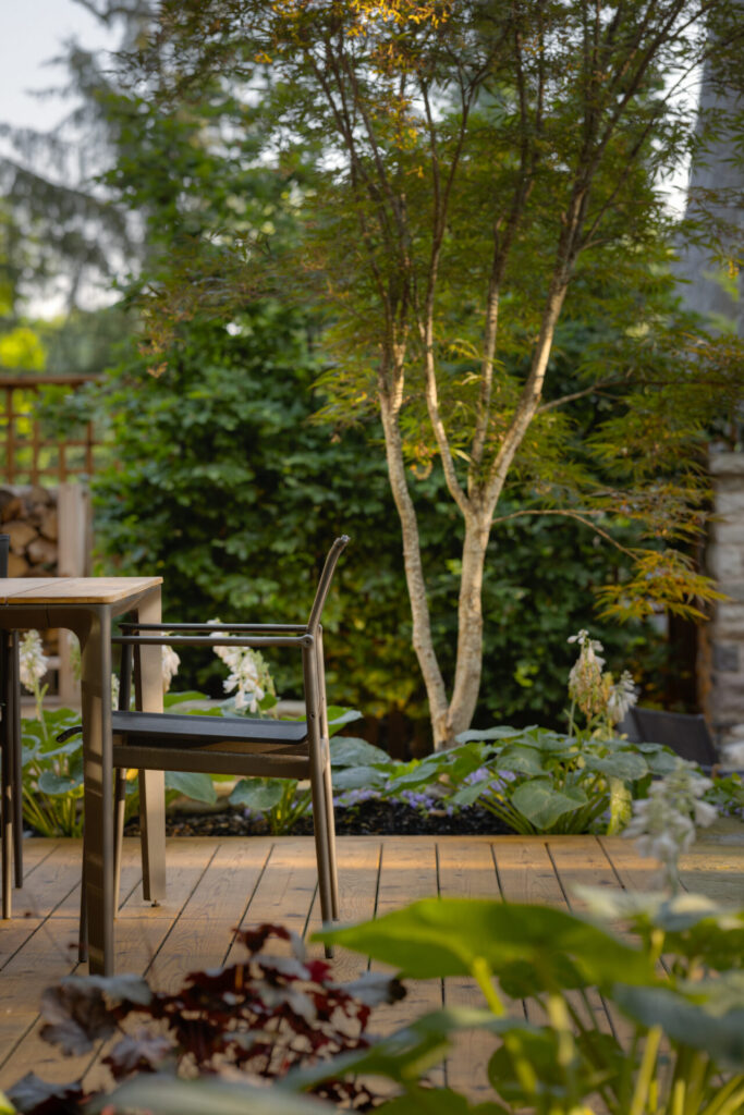 A serene garden space with a wooden deck, modern outdoor furniture, lush greenery, and trees, creating a peaceful atmosphere for relaxation or contemplation.