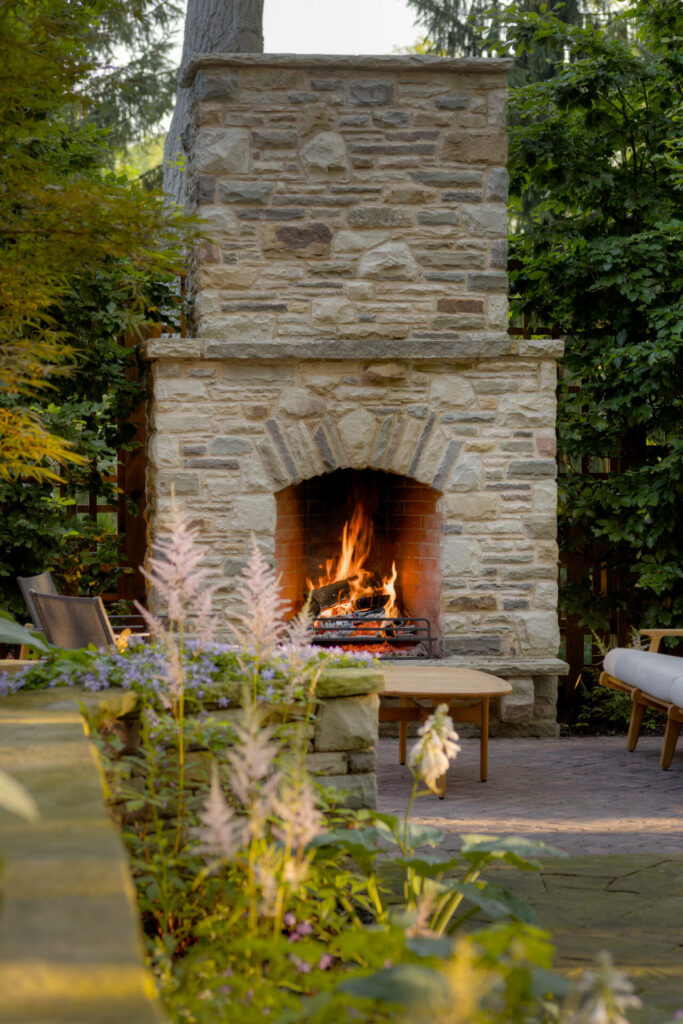 An outdoor stone fireplace is alight with a cozy fire, surrounded by lush greenery, flowers, and comfortable patio furniture, creating a serene backyard setting.