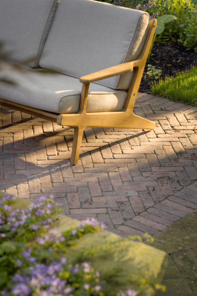 This image features a cozy outdoor setting with a modern, gray cushioned sofa and a wooden armrest visible, set on a brick patio surrounded by greenery and flowers.