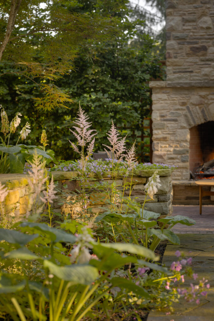 A serene garden scene with flowering plants in the foreground and a stone fireplace in the background, surrounded by lush greenery and trees.