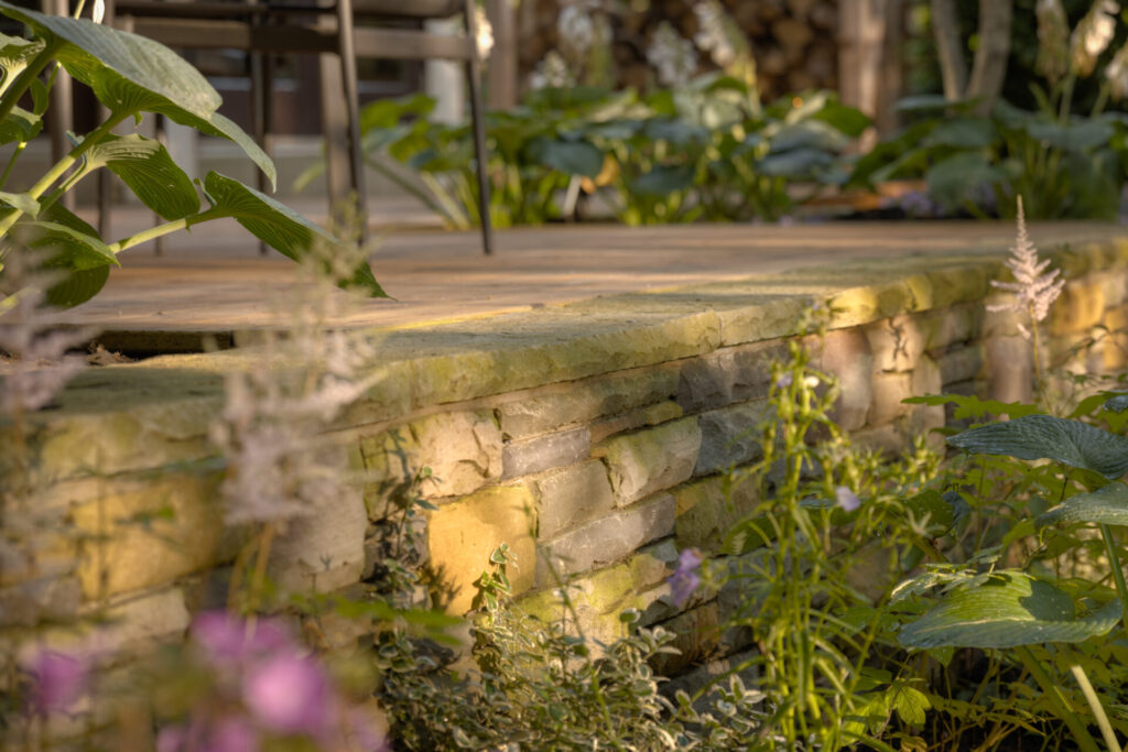 The image shows a part of a garden with a stone wall, blurred foliage in the foreground, a wooden deck, and patio chairs in the background.