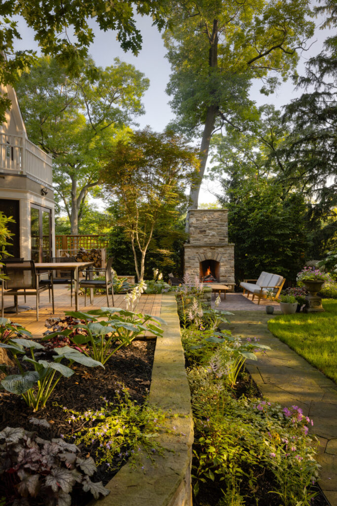 A serene backyard garden with a stone patio, outdoor fireplace, lush landscaping, dining furniture, and surrounding tall trees in a tranquil setting.