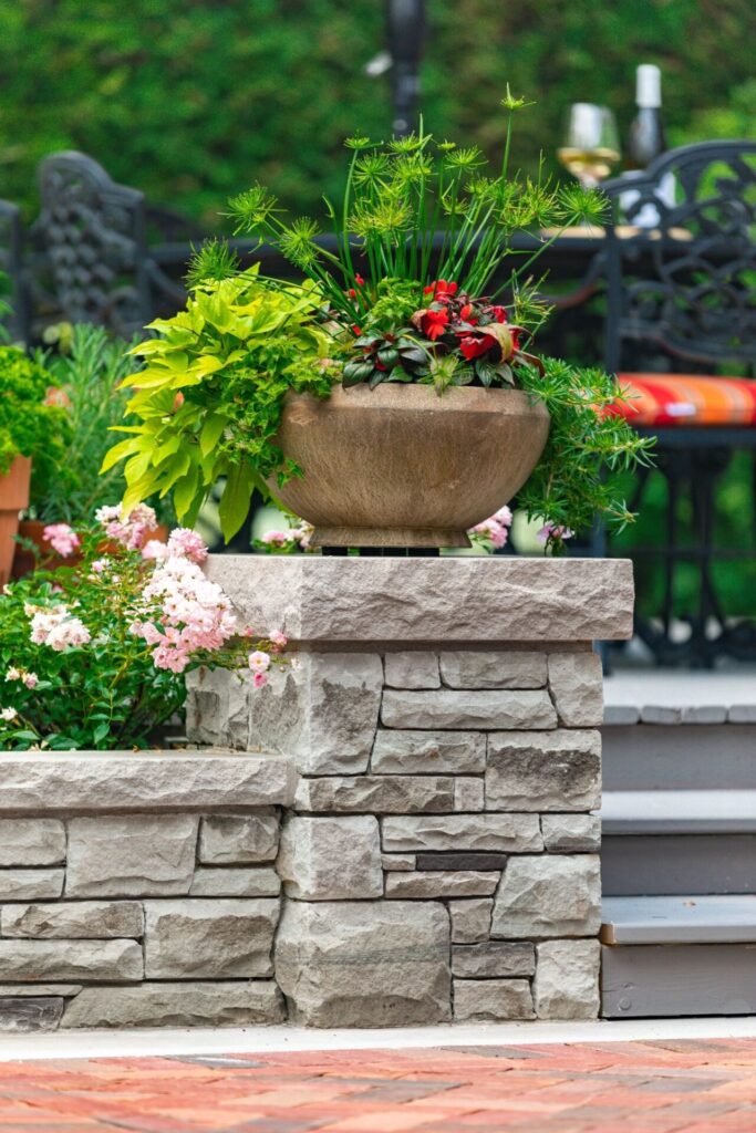 A colorful planter sits atop a stone pillar, surrounded by lush greenery, with patio furniture and pink flowers in the background.